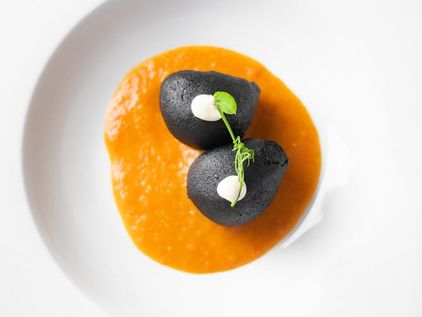 Dos croquetas negras sobre una salsa anaranjada, con mahonesa y un brote verde